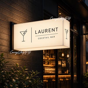以下が含まれることがあります： 黒いフレームの「Laurent Cocktail Bar」の長方形の電光掲示板。看板には、すっきりとしたフォントで「Cocktail Bar」の上に「Laurent」という名前が表示され、両側にカクテルグラスのアイコンが付いています。看板は暗いレンガの壁に取り付けられています。