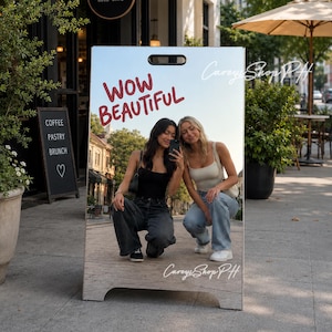 Może przedstawiać: Dwustronny szyld w kształcie litery A z napisem "WOW BEAUTIFUL" w czerwonym skrypcie. Szyld przedstawia zdjęcie dwóch kobiet robiących sobie selfie. Szyld znajduje się przed kawiarnią z menu na tablicy.