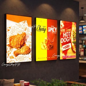 Puede incluir: Tres carteles de comida enmarcados en una pared gris oscuro. Un cartel presenta pollo frito sobre papel estampado. El segundo cartel dice "Cheesy Spicy Chicken". El tercer cartel dice "Hot Dog?" con un precio.