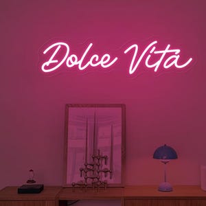 May include: A neon sign displaying the words "Dolce Vita" in a cursive font, illuminated in bright pink. The sign is mounted on a pink wall above a wooden cabinet with decorative items, including a mirror, books, and a small lamp.