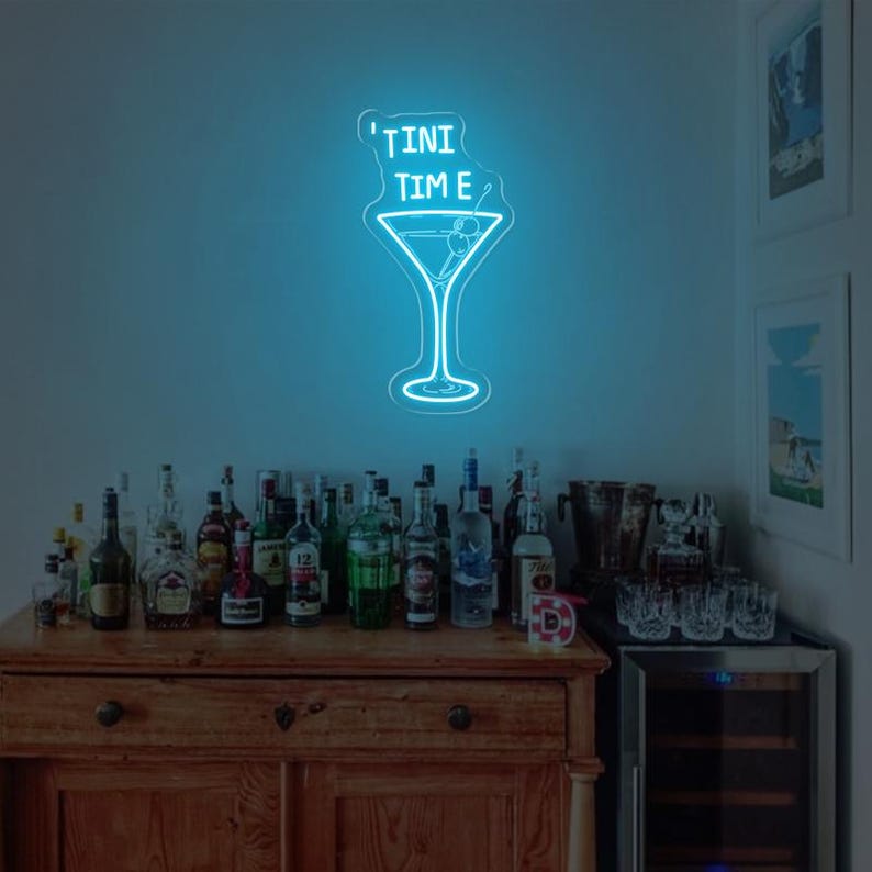 May include: A neon sign featuring a martini glass and the text "'TINI TIME" in blue. The sign is mounted on a wall above a bar cart stocked with bottles of spirits and glasses.