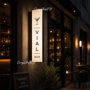 Pode incluir: Uma placa retangular iluminada para "VIAL BAR" com um &iacute;cone de copo de coquetel, montada em uma parede de pedra. A entrada do bar &eacute; vis&iacute;vel, e uma lousa abaixo diz "COCKTAILS, WINE, GOOD TIMES."