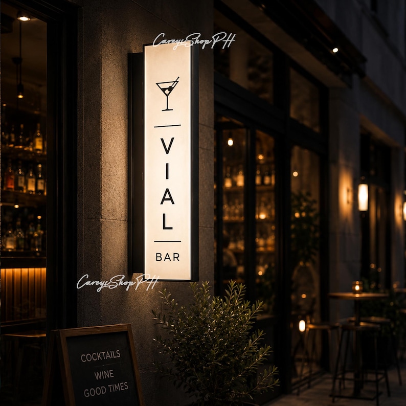 Pode incluir: Uma placa retangular iluminada para "VIAL BAR" com um &iacute;cone de copo de coquetel, montada em uma parede de pedra. A entrada do bar &eacute; vis&iacute;vel, e uma lousa abaixo diz "COCKTAILS, WINE, GOOD TIMES."