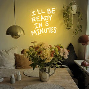 Może przedstawiać: Scena w jadalni z neonowym znakiem z napisem "I'LL BE READY IN 5 MINUTES". Bukiet kolorowych kwiat&oacute;w w srebrnym dzbanku stoi na drewnianym stole. Widoczna jest r&oacute;wnież złota lampa wisząca i mała lampa.