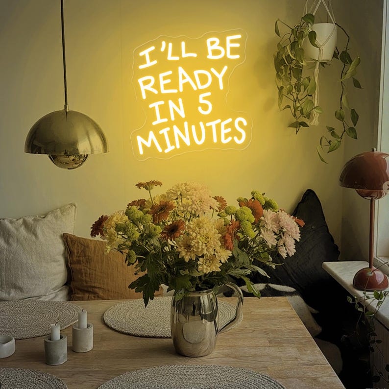 Może przedstawiać: Scena w jadalni z neonowym znakiem z napisem "I'LL BE READY IN 5 MINUTES". Bukiet kolorowych kwiat&oacute;w w srebrnym dzbanku stoi na drewnianym stole. Widoczna jest r&oacute;wnież złota lampa wisząca i mała lampa.