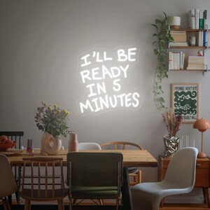 Może przedstawiać: Biały neonowy znak z napisem "I'LL BE READY IN 5 MINUTES" na ścianie. Znak znajduje się nad drewnianym stołem jadalnym z krzesłami. Widoczne są r&oacute;wnież elementy dekoracyjne, w tym kwiaty i plakat.