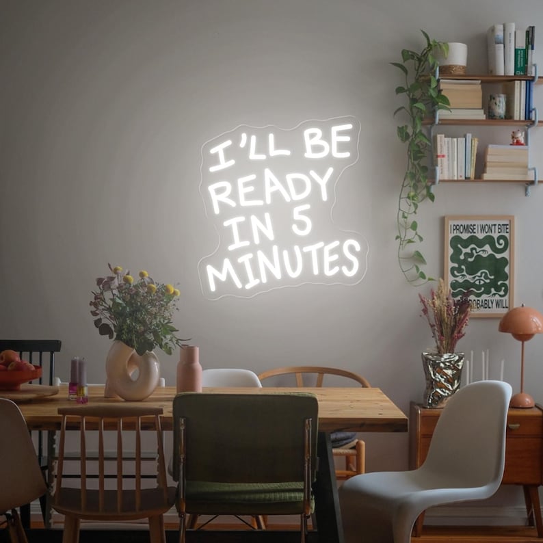 Może przedstawiać: Biały neonowy znak z napisem "I'LL BE READY IN 5 MINUTES" na ścianie. Znak znajduje się nad drewnianym stołem jadalnym z krzesłami. Widoczne są r&oacute;wnież elementy dekoracyjne, w tym kwiaty i plakat.