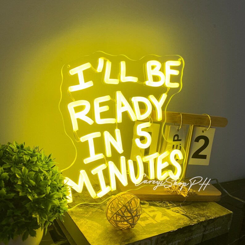 Może przedstawiać: Ż&oacute;łty neon z napisem "I'LL BE READY IN 5 MINUTES". Znak znajduje się na drewnianej podstawie z kalendarzem pokazującym "SEP 2". Widoczna jest r&oacute;wnież mała dekoracyjna kulka i roślina doniczkowa.