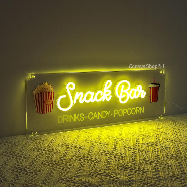 May include: A yellow neon sign with the words "Snack Bar" in a cursive font. The sign also includes a red and white striped popcorn bucket and a red cup with a straw. The text below the neon sign reads "Drinks - Candy - Popcorn".
