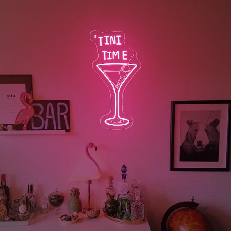 May include: Pink neon sign with a martini glass and the text "'TINI TIME" hanging on a pink wall. The sign is lit up and there are other decorations in the background.