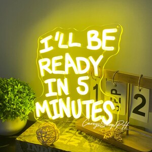Może przedstawiać: Ż&oacute;łty neon z napisem "I'LL BE READY IN 5 MINUTES" w kolorze białym. Znak znajduje się na drewnianym stojaku z kalendarzem pokazującym datę. Widoczna jest r&oacute;wnież mała roślina i dekoracyjna kula.