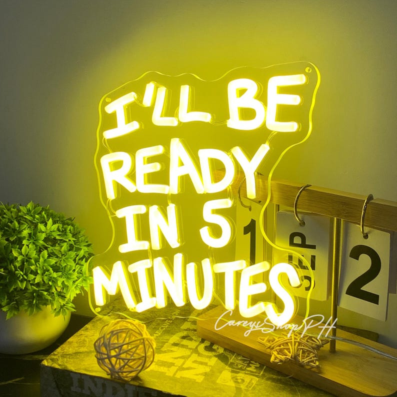 Może przedstawiać: Ż&oacute;łty neon z napisem "I'LL BE READY IN 5 MINUTES" w kolorze białym. Znak znajduje się na drewnianym stojaku z kalendarzem pokazującym datę. Widoczna jest r&oacute;wnież mała roślina i dekoracyjna kula.