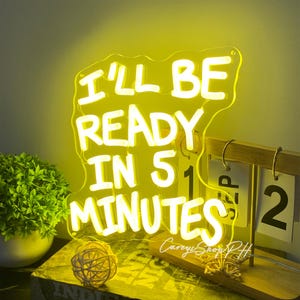Może przedstawiać: Ż&oacute;łty neon z napisem "I'LL BE READY IN 5 MINUTES". Znak jest zamontowany na przezroczystym akrylowym podkładzie. Mała roślina doniczkowa, drewniany kalendarz i dekoracyjna kula znajdują się na pierwszym planie.