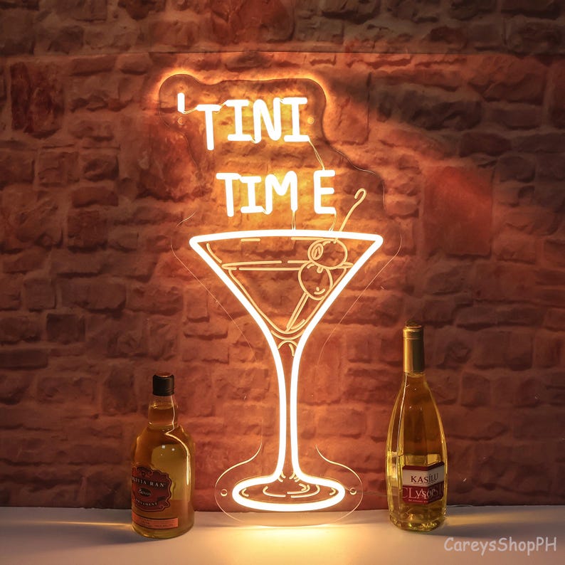 May include: Neon sign featuring a martini glass and the text "TINI TIME" in white. Two bottles of wine are positioned on either side of the sign.