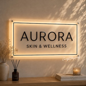Può includere: Un'insegna rettangolare in acrilico montata a parete con il testo nero AURORA SKIN & WELLNESS. L'insegna è retroilluminata con una luce calda, incorniciata da un sottile bordo nero e fissata alla parete con quattro distanziatori in metallo.