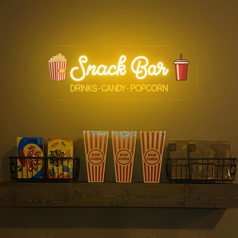 May include: A yellow neon sign that reads "Snack Bar" with a red and white popcorn bucket and a red cup with a straw on either side. The sign also says "Drinks - Candy - Popcorn". Below the sign are three red and white striped popcorn containers and two bags of snacks on a wooden shelf.
