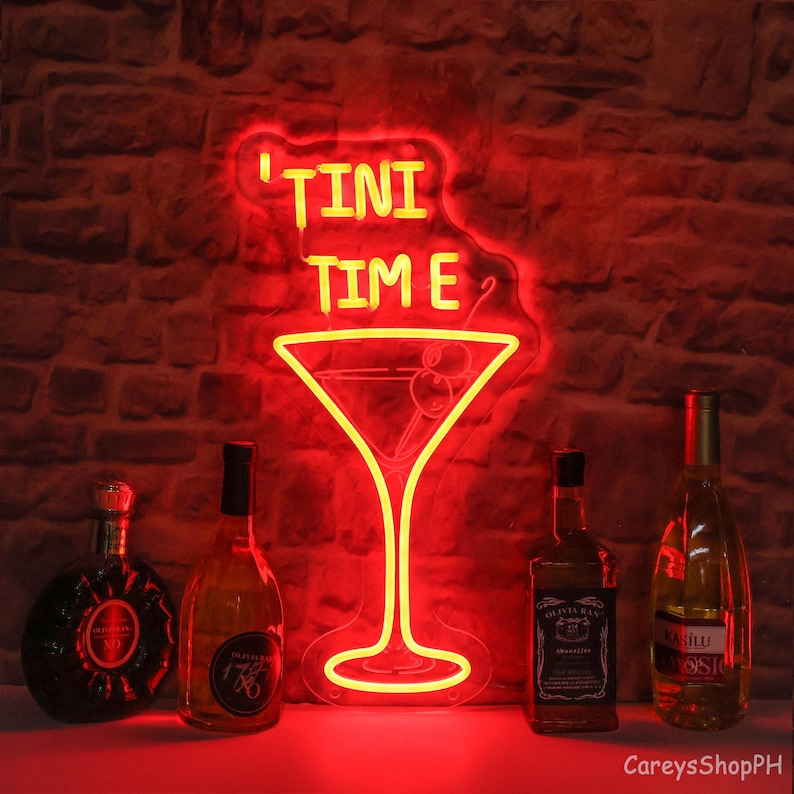 May include: Red neon sign with the text "Tini Time" and a martini glass shape. The sign is hanging on a brick wall with four bottles of liquor in front of it.