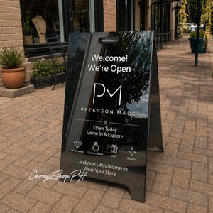 Puede incluir: Un letrero negro en forma de A con el texto "Welcome! We're Open" y "PM PETERSON MADE." El letrero también dice "Open Today Come In & Explore" y enumera joyería fina, diseño personalizado, regalos y reparaciones. El letrero está sobre una acera de ladrillos.