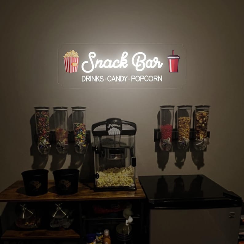 May include: A snack bar setup with a neon sign that reads "Snack Bar Drinks - Candy - Popcorn". There are three dispensers filled with candy, a popcorn machine, and two buckets.
