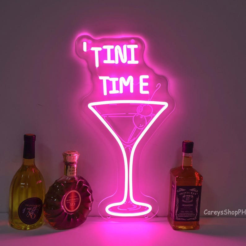 May include: Pink neon sign with the text "'Tini Time" and a martini glass with olives. The sign is hanging on a white wall with three bottles of liquor in front of it.