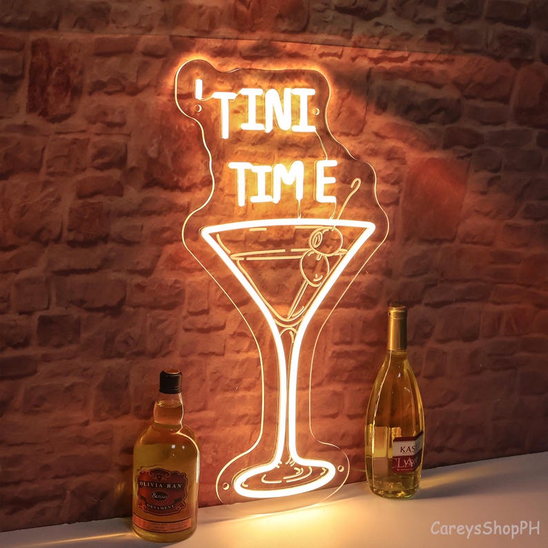 May include: Neon sign featuring a martini glass and the text "TINI TIME" in white. The sign is lit with a warm yellow glow. Two bottles of spirits are in the foreground.