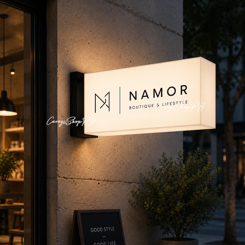 Pode incluir: Uma placa retangular iluminada para "NAMOR BOUTIQUE & LIFESTYLE" est&aacute; montada na fachada de um edif&iacute;cio. A placa &eacute; branca com letras pretas e um logotipo minimalista. Uma pequena placa de quadro-negro abaixo diz "GOOD STYLE GOOD LIFE".