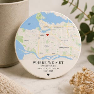 Puede incluir: Un posavasos redondo y blanquecino con un mapa de Vancouver, BC, y un corazón rojo que marca un lugar. El texto dice "WHERE WE MET" e incluye coordenadas y una fecha. Un pequeño corazón negro en la parte inferior.