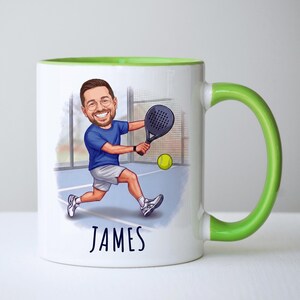 Puede incluir: Taza de cer&aacute;mica blanca con asa y borde verdes, con una ilustraci&oacute;n de dibujos animados de una persona jugando al p&aacute;del. La persona lleva ropa deportiva azul y blanca, sosteniendo una raqueta y una pelota amarilla. El nombre "JAMES" est&aacute; escrito debajo.
