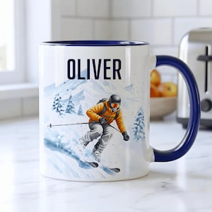 May include: White ceramic mug with a blue handle and rim, featuring a skier in orange and gray ski gear on a snowy mountain scene. The name "OLIVER" is printed in black on the mug.