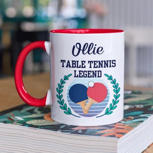 May include: White ceramic mug with a red interior and handle. The mug features the text "Ollie TABLE TENNIS LEGEND" above a graphic of two ping pong paddles and a ball, surrounded by a green wreath.