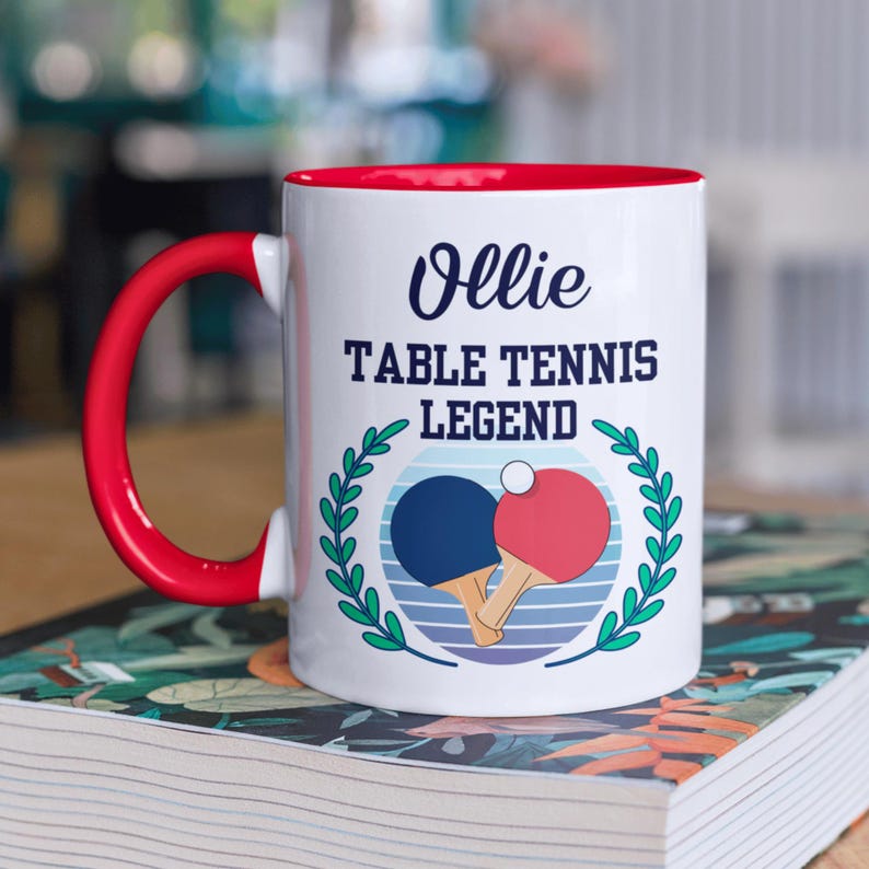 May include: White ceramic mug with a red interior and handle. The mug features the text "Ollie TABLE TENNIS LEGEND" above a graphic of two ping pong paddles and a ball, surrounded by a green wreath.