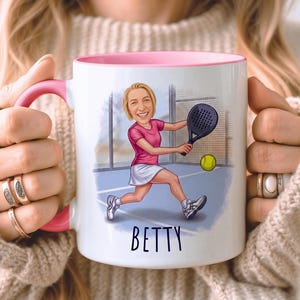 Puede incluir: Taza de cer&aacute;mica blanca con interior y asa rosas. La taza presenta una ilustraci&oacute;n de dibujos animados de una mujer jugando al p&aacute;del, con el nombre "BETTY" en escritura negra. La mujer lleva una camiseta rosa y una falda blanca.