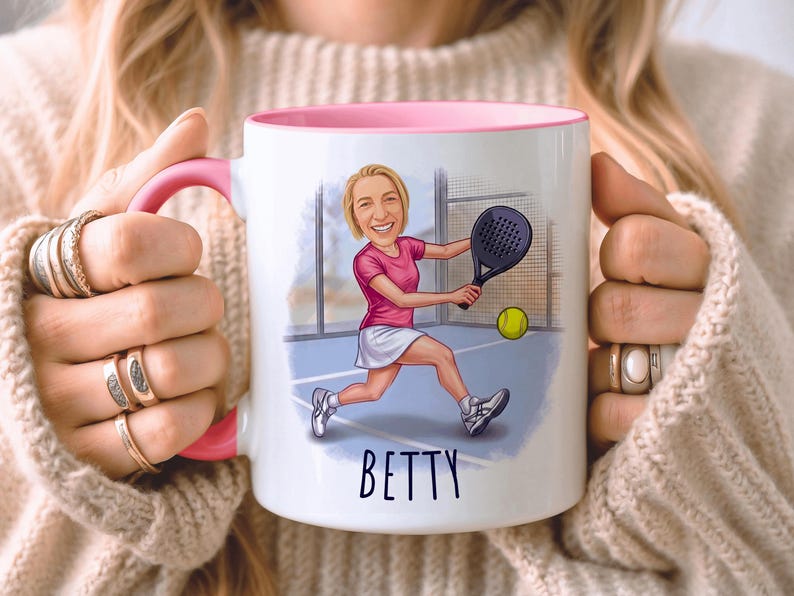 Puede incluir: Taza de cer&aacute;mica blanca con interior y asa rosas. La taza presenta una ilustraci&oacute;n de dibujos animados de una mujer jugando al p&aacute;del, con el nombre "BETTY" en escritura negra. La mujer lleva una camiseta rosa y una falda blanca.