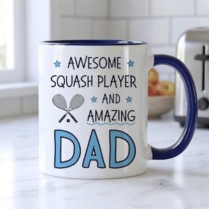 May include: White ceramic mug with a blue interior and handle. The mug features the text "AWESOME SQUASH PLAYER AND AMAZING DAD" in blue, along with squash rackets and a ball graphic. A great gift for a squash-playing dad.