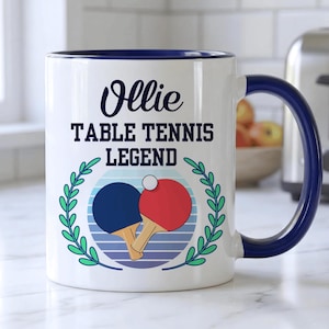 May include: White ceramic mug with a blue handle and rim. The mug features the name "Ollie" above the text "TABLE TENNIS LEGEND". A graphic of two ping pong paddles and a ball is in the center, surrounded by a green wreath.