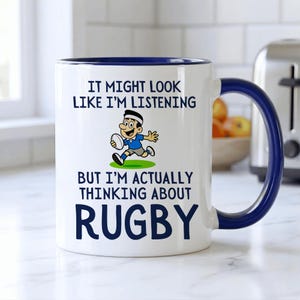 Può includere: Tazza in ceramica bianca con manico e bordo blu. La tazza presenta il testo "IT MIGHT LOOK LIKE I'M LISTENING BUT I'M ACTUALLY THINKING ABOUT RUGBY" e un giocatore di rugby dei cartoni animati.