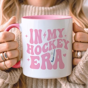 May include: A white ceramic mug with a pink interior and a pink handle. The front of the mug features the pink text "IN MY HOCKEY ERA" in a wavy, retro-style font, accompanied by small pink star, lightning bolt, heart, hockey stick, and puck icons.