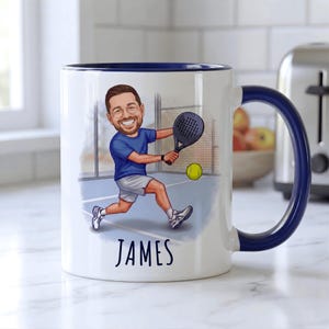 Puede incluir: Taza de cer&aacute;mica blanca con asa y borde azules, con una ilustraci&oacute;n de dibujos animados de una persona jugando al p&aacute;del. La ilustraci&oacute;n incluye una cancha de p&aacute;del, una pelota amarilla y el nombre "JAMES" en negro.