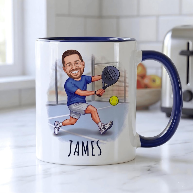 Puede incluir: Taza de cer&aacute;mica blanca con asa y borde azules, con una ilustraci&oacute;n de dibujos animados de una persona jugando al p&aacute;del. La ilustraci&oacute;n incluye una cancha de p&aacute;del, una pelota amarilla y el nombre "JAMES" en negro.