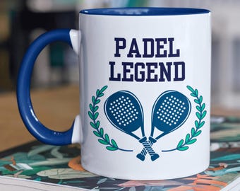 Puede incluir: Taza de cerámica blanca con borde y asa azules. La taza tiene un gráfico de dos raquetas de pádel cruzadas con hojas verdes y el texto "PADEL LEGEND" en azul.