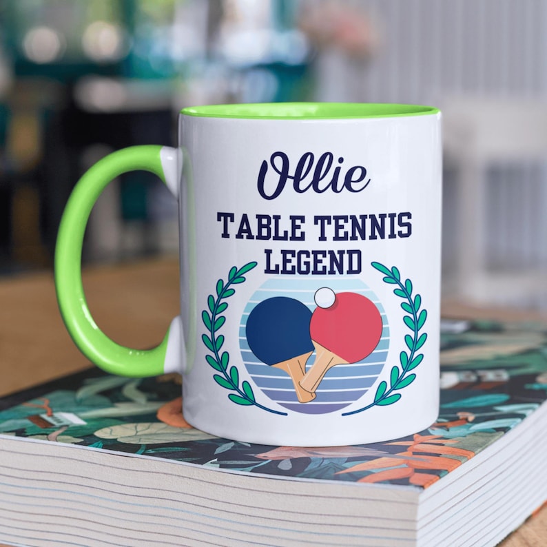 May include: White ceramic mug with a green handle and interior. The mug features the text "Ollie TABLE TENNIS LEGEND" above a graphic of two ping pong paddles and a ball, surrounded by a green laurel wreath.