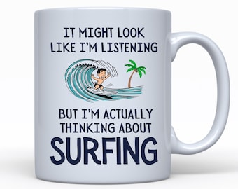 Könnte beinhalten: Eine weiße Keramik-Kaffeetasse mit einer blauen und weißen Cartoon-Illustration eines Surfers, der auf einer Welle reitet. Der Text auf der Tasse lautet "It might look like I'm listening but I'm actually thinking about surfing."