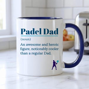 May include: White ceramic mug with a blue rim and handle. The mug features the text "Padel Dad" in blue, along with a definition and a silhouette of a person playing padel. A great gift for a padel-playing dad.