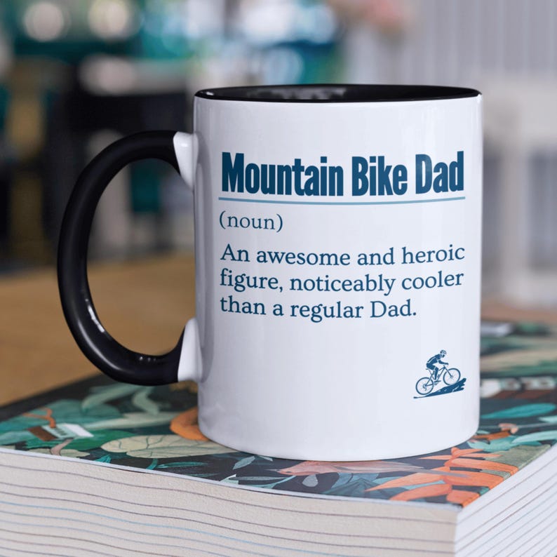 May include: White ceramic mug with a black rim and handle. The mug has a blue and white design that reads "Mountain Bike Dad (noun) An awesome and heroic figure, noticeably cooler than a regular Dad."  A small blue and white illustration of a person riding a mountain bike is at the bottom of the mug.