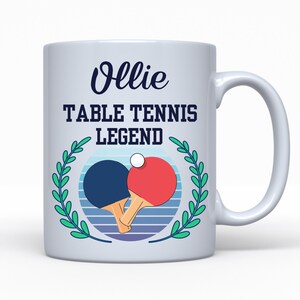 May include: White ceramic mug with a handle, featuring the name "Ollie" in a script font above the text "TABLE TENNIS LEGEND." A blue and red ping pong paddle design is centered, surrounded by a green laurel wreath.