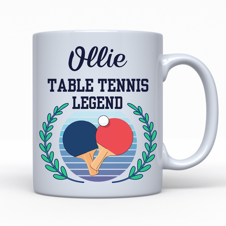 May include: White ceramic mug with a handle, featuring the name "Ollie" in a script font above the text "TABLE TENNIS LEGEND." A blue and red ping pong paddle design is centered, surrounded by a green laurel wreath.