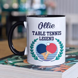 May include: White ceramic mug with a black handle and rim. The mug features the text "Ollie TABLE TENNIS LEGEND" in navy blue, along with a graphic of two ping pong paddles and a ball, surrounded by a green wreath.