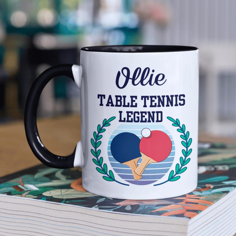 May include: White ceramic mug with a black handle and rim. The mug features the text "Ollie TABLE TENNIS LEGEND" in navy blue, along with a graphic of two ping pong paddles and a ball, surrounded by a green wreath.