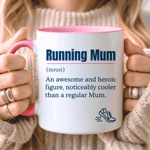 May include: A white ceramic mug with a pink interior and handle. The front features dark blue text that reads Running Mum (noun) An awesome and heroic figure, noticeably cooler than a regular Mum. A small dark blue graphic of a running shoe is below.