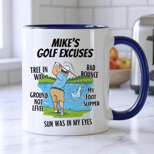 May include: White ceramic mug with a blue interior and handle, featuring a golf-themed design. The mug has the text "MIKE'S GOLF EXCUSES" and illustrations of a golfer and golf course. Additional text includes "TREE IN WAY", "GROUND NOT LEVEL", and "SUN WAS IN MY EYES".